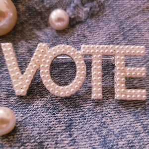 May include: A brooch spelling the word VOTE in capital letters, constructed from a silver-toned metal frame densely covered in small, round, cream-colored faux pearls. The piece is displayed alongside three loose, spherical faux pearls.