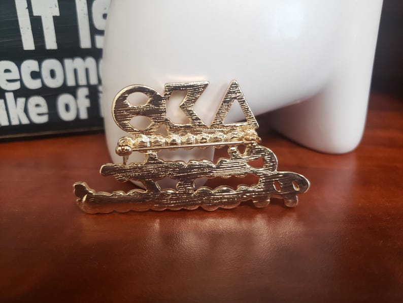 May include: A gold-colored pin with the Greek letters Omega, Beta, and Alpha, with the word "Sisterhood" below. The pin is likely a sorority or organization emblem, with a decorative design.