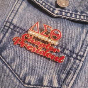 May include: A red and gold enamel pin featuring the Greek letters Delta, Sigma, and Theta, and the words "Chapter President." The pin is decorated with faux pearls and is set against a blue denim jacket.