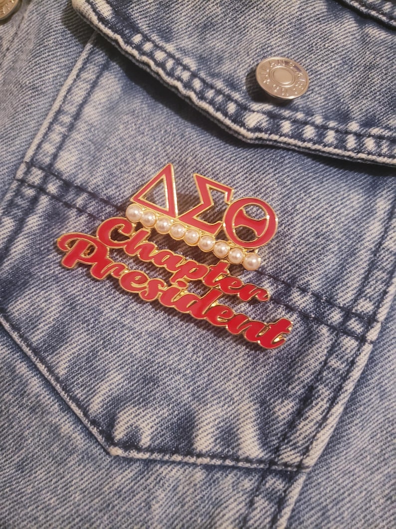 May include: A red and gold enamel pin featuring the Greek letters Delta, Sigma, and Theta, and the words "Chapter President." The pin is decorated with faux pearls and is set against a blue denim jacket.