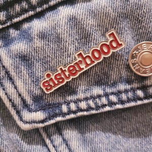 May include: A red and white enamel pin with the word "sisterhood" in a decorative font. The pin is attached to a denim jacket. A silver button is visible to the right of the pin.