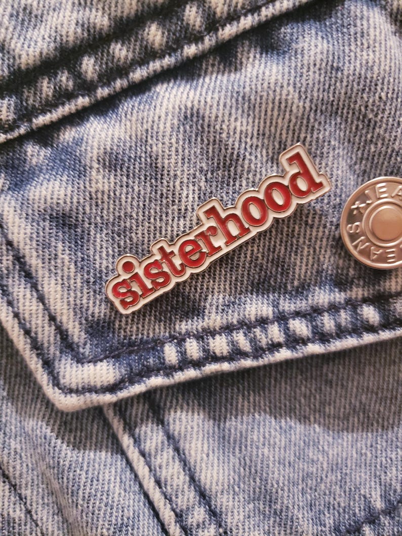 May include: A red and white enamel pin with the word "sisterhood" in a decorative font. The pin is attached to a denim jacket. A silver button is visible to the right of the pin.