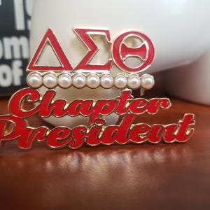 May include: A gold-toned pin with red enamel lettering that reads "Chapter President." Above the text are three Greek letters in red enamel, with a row of pearls. The pin is set against a blurred background.