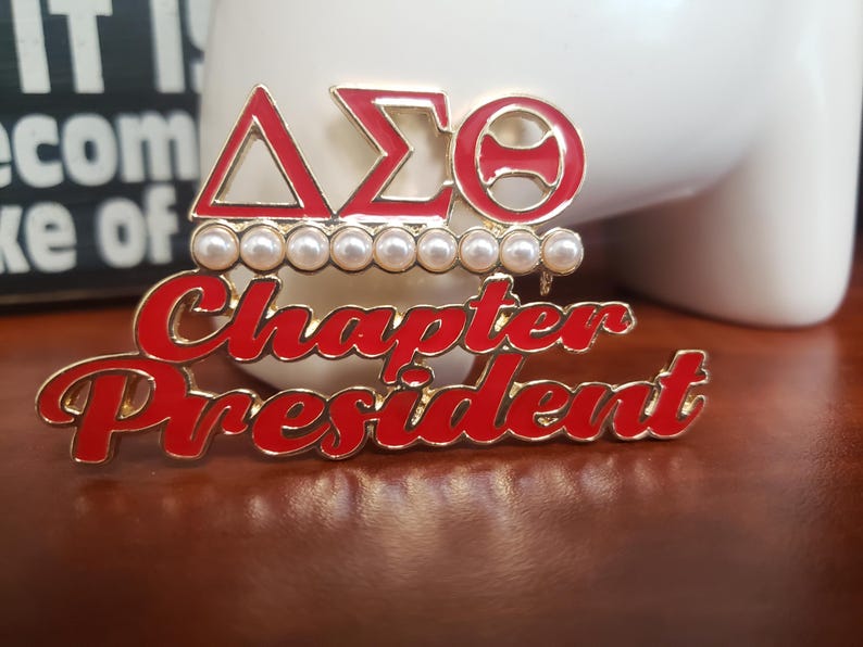 May include: A gold-toned pin with red enamel lettering that reads "Chapter President." Above the text are three Greek letters in red enamel, with a row of pearls. The pin is set against a blurred background.