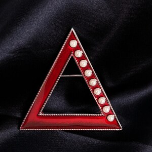 May include: A red enamel triangle brooch with a silver-colored border. The brooch is adorned with a row of white pearls along one side. The brooch is set against a black background.