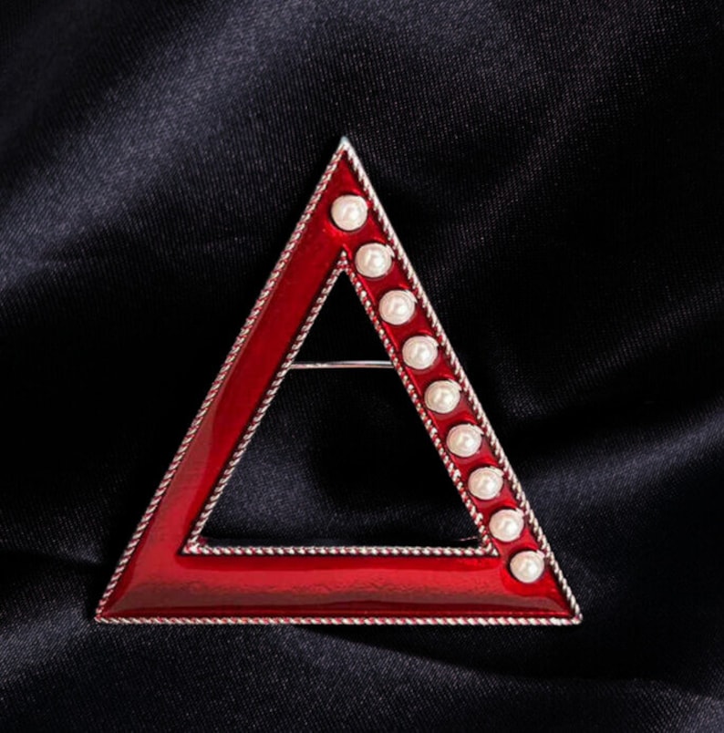 May include: A red enamel triangle brooch with a silver-colored border. The brooch is adorned with a row of white pearls along one side. The brooch is set against a black background.