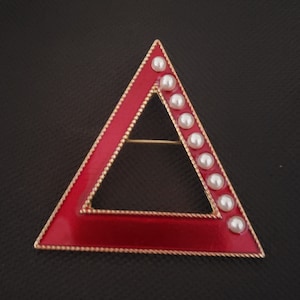 May include: A red enamel triangle brooch with a gold-colored border and a row of white faux pearls along one side. The brooch has a pin on the back for secure fastening. The jewelry is set against a dark background.
