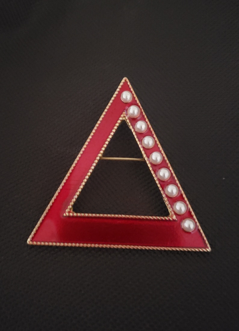 May include: A red enamel triangle brooch with a gold-colored border and a row of white faux pearls along one side. The brooch has a pin on the back for secure fastening. The jewelry is set against a dark background.