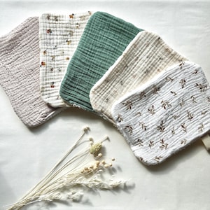 May include: A collection of five soft, textured baby washcloths in various colors and patterns. The cloths include solid light pink, floral print, solid green, cream with floral print, and white with brown leaf print. A bundle of dried flowers is in the foreground.