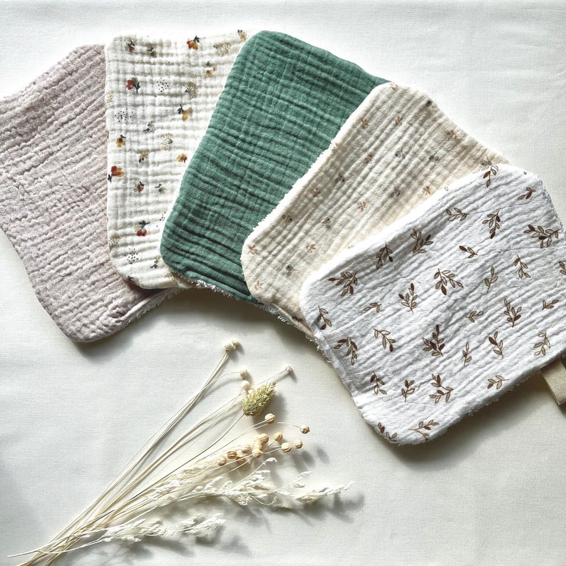 May include: A collection of five soft, textured baby washcloths in various colors and patterns. The cloths include solid light pink, floral print, solid green, cream with floral print, and white with brown leaf print. A bundle of dried flowers is in the foreground.