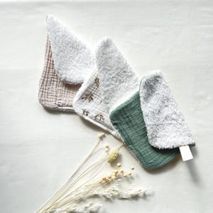 May include: A collection of baby bibs in various colors and textures. The bibs are in shades of pink, white, and green, with a soft, textured appearance. Some bibs feature leaf patterns. The bibs are arranged on a white surface with dried flowers.