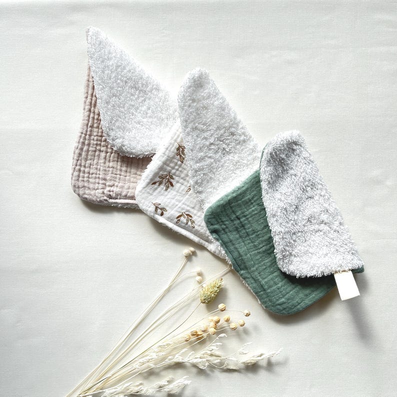 May include: A collection of baby bibs in various colors and textures. The bibs are in shades of pink, white, and green, with a soft, textured appearance. Some bibs feature leaf patterns. The bibs are arranged on a white surface with dried flowers.