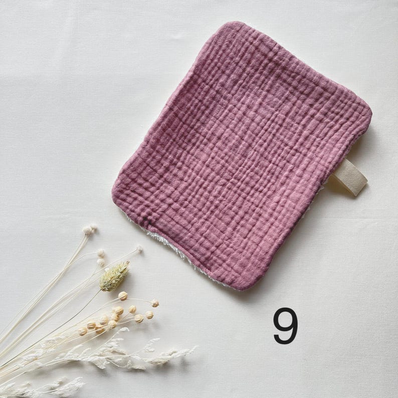 May include: A dusty rose-colored, square baby washcloth with a textured, crinkled surface. It has a cream-colored loop on one corner and a white, fluffy trim. The number "9" is printed in black.