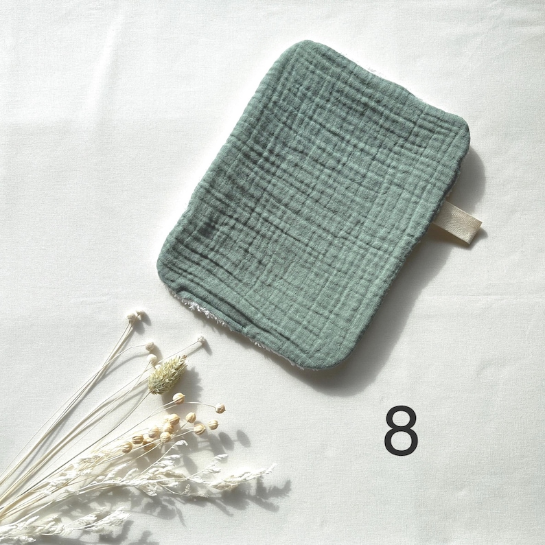 May include: A sage green washcloth with a textured, crinkled appearance. The washcloth has a small loop on one side and is set against a white background. The number "8" is printed in black on the lower right corner.