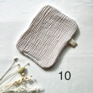 May include: A light beige, rectangular washcloth with a textured, crinkled surface. A small, light beige fabric loop is attached to one corner. The number "10" is printed in black on the bottom right corner. Dried flowers are in the bottom left corner.