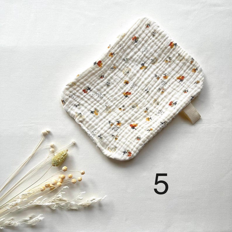 May include: A white, rectangular baby burp cloth with a floral pattern in orange, yellow, and black. The cloth has a textured, quilted appearance and a small loop on one corner. The number "5" is printed in black.