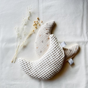 Könnte beinhalten: Zwei halbmondförmige Babykissen. Eines ist weiß mit Waffelstruktur, das andere beige mit floralem Muster. Beide haben kleine Etiketten mit der Aufschrift "EVA" und "OLD". Die Kissen liegen auf einer weißen Oberfläche mit getrockneten Blumen.