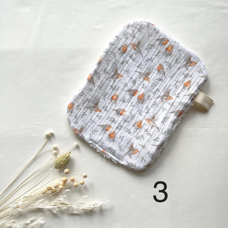 May include: A white and gray washcloth with a bird and leaf pattern. The washcloth has a loop and is decorated with orange birds and gray leaves. The number 3 is printed in black on the bottom right.