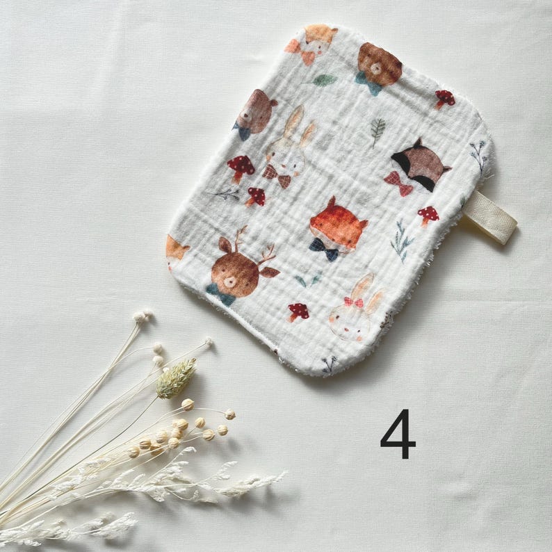 May include: White baby washcloth with a whimsical forest animal print. The washcloth features illustrations of deer, foxes, bears, and rabbits, along with red mushrooms and green leaves. A cream-colored loop is attached to the edge. The number "4" is printed in black.