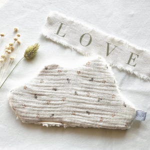 Peut inclure: Un article textile en forme de nuage avec un tissu beige à motif floral sur une face et un tissu blanc texturé sur l'autre. Une bande de tissu effilochée avec le mot LOVE imprimé en vert et une petite étiquette marquée EVA sont visibles.