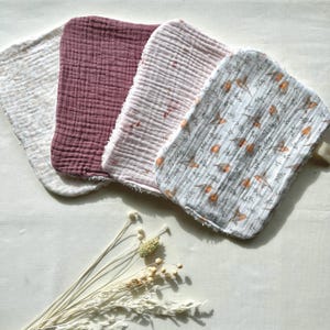 May include: Four soft, textured washcloths in varying colors: off-white, deep rose, light pink, and a patterned cloth with birds and branches. The washcloths are arranged in a row, with a neutral background and dried flowers.