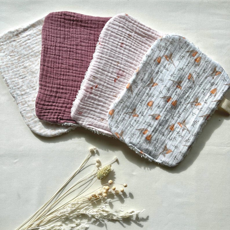 May include: Four soft, textured washcloths in varying colors: off-white, deep rose, light pink, and a patterned cloth with birds and branches. The washcloths are arranged in a row, with a neutral background and dried flowers.
