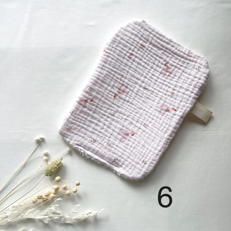 May include: A light pink, rectangular baby washcloth with a floral pattern. The washcloth has a textured, crinkled appearance and a small loop on one end. The number "6" is printed in black on the bottom right.