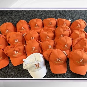 May include: A collection of 18 orange baseball caps with white embroidered letters and the text "Willy's Hats" on each cap. One white baseball cap with the letter "H" and the text "Get On It" is in the center of the image.