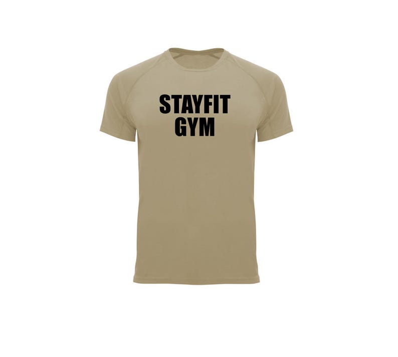 May include: A tan short-sleeved athletic shirt with the text "STAYFIT GYM" in bold black letters across the chest. The shirt has a crew neck and raglan sleeves. The background is plain white.