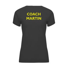 Op de afbeelding: Een donkergrijs t-shirt met de tekst "COACH MARTIN" in gele blokletters op de achterkant. Het shirt heeft korte mouwen en een V-hals.