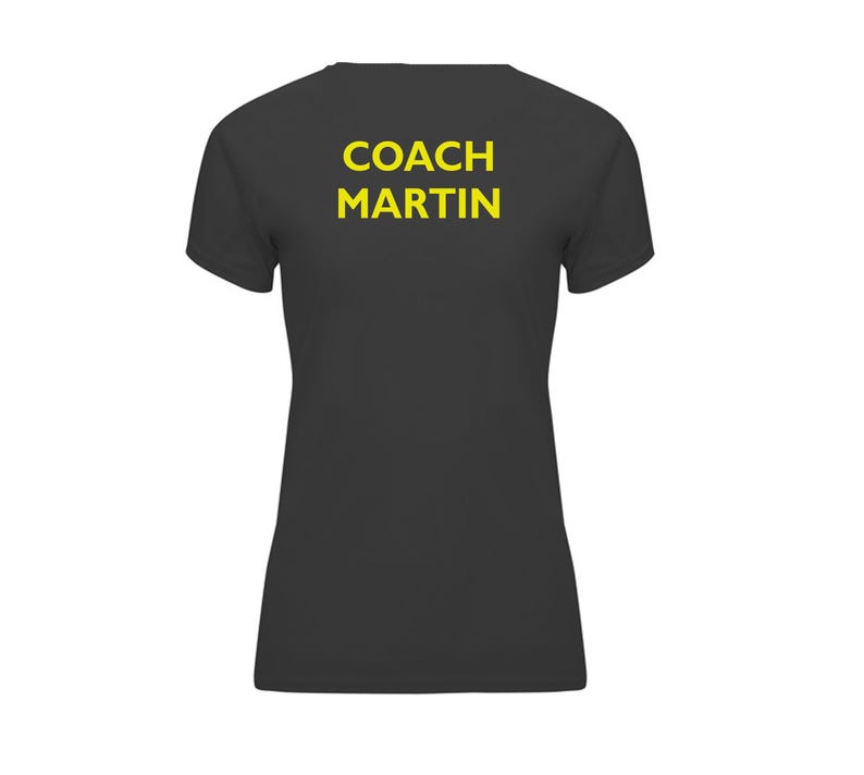 Op de afbeelding: Een donkergrijs t-shirt met de tekst "COACH MARTIN" in gele blokletters op de achterkant. Het shirt heeft korte mouwen en een V-hals.