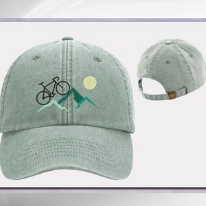 May include: A light green, adjustable, cotton baseball cap with an embroidered design of a bicycle riding up a mountain with a sun in the background.