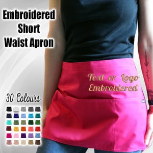 May include: A pink short waist apron with two pockets and a gold embroidered text that says "Text or Logo Embroidered". The apron is shown being worn by a person. There are 30 different colors of aprons shown in the image.