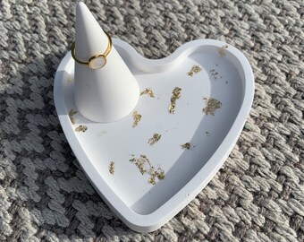 May include: White heart-shaped tray with gold flecks and a white ring holder cone. The tray is made of resin and has a smooth, glossy finish.