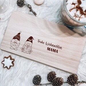 May include: A wooden chopping board with a Christmas gnome design and the text "Frohe Weihnachten MAMA". The board is surrounded by Christmas decorations, including a mug of hot chocolate with marshmallows, gingerbread cookies, and pine branches.