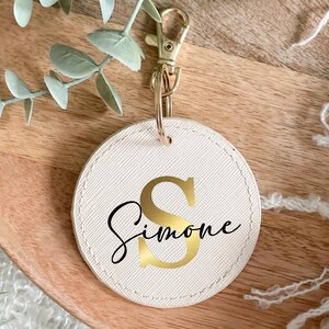 Könnte beinhalten: Ein weißer Leder-Schlüsselanhänger mit einem goldenen Buchstaben "S" und dem Namen "Simone" in goldener Schrift.