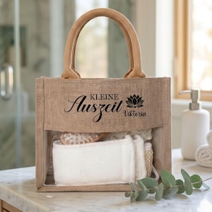 May include: A natural burlap tote bag with a clear window, filled with bath accessories. The bag has a woven handle and features the text "Kleine Auszeit Viktoria" in black script. Inside are white washcloths and a wooden brush.