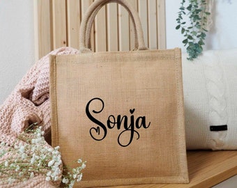 May include: A brown burlap tote bag with a black cursive name "Sonja" and a small heart above the "a".