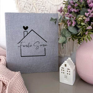 Könnte beinhalten: Graues Fotoalbum mit einem Hausdesign und dem Text "Familie Simon" in schwarzer Schrift. Im Hintergrund befinden sich eine kleine weiße Hausfigur und eine Vase mit lila Blumen.