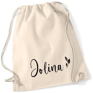 Könnte beinhalten: Cremefarbener Turnbeutel mit dem Namen "Jolina" in schwarzer Schrift, begleitet von einem kleinen Blattdesign. Die Tasche hat ein schlichtes, lässiges Design mit Kordelzug und neutraler Farbe.