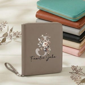 Peut inclure: Un portefeuille zippé taupe avec un motif floral et la lettre "J". Le portefeuille a une dragonne et le texte "Familie Jahn". D'autres portefeuilles en turquoise, marron, gris, noir, beige et rose sont empilés en arrière-plan.
