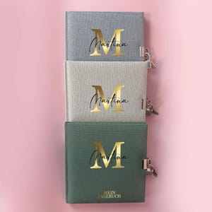 Może przedstawiać: Trzy zamknięte dzienniki z pozłacanym inicjałem "M" i imieniem "Martina" na okładce. Dzienniki są ułożone jeden na drugim, przy czym górny dziennik jest szary, środkowy beżowy, a dolny zielony. Na dolnym dzienniku nadrukowany jest tekst "MEIN TAGEBUCH".