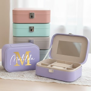Könnte beinhalten: Eine Ansammlung pastellfarbener Schmuckkästchen, darunter rosa, mintgrün und grau, mit einer personalisierten lila Schatulle im Vordergrund. Die offene lila Schatulle zeigt einen Spiegel und Fächer. Der Text "Matilda" ist auf der Vorderseite zu sehen.