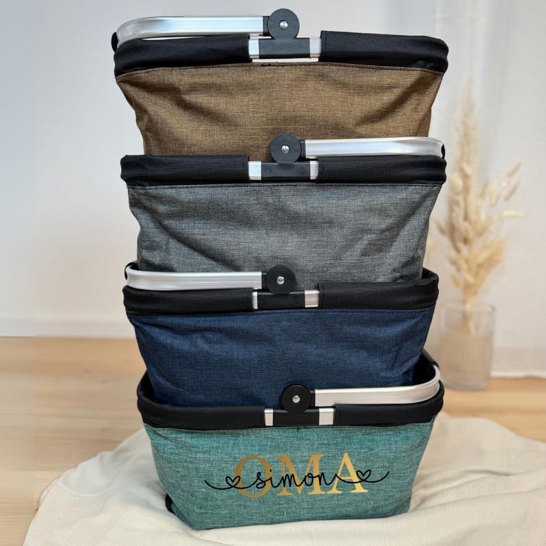 May include: Four rectangular fabric baskets stacked on top of each other, each with a silver handle and black trim. The baskets are in shades of brown, grey, blue, and teal. The bottom teal basket has the gold text "OMA Simon".