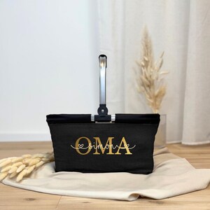 May include: A black tote bag with a silver handle and the word "OMA" in gold script. Below "OMA" is the name "simone" in a cursive font. The bag is sitting on a beige cloth.