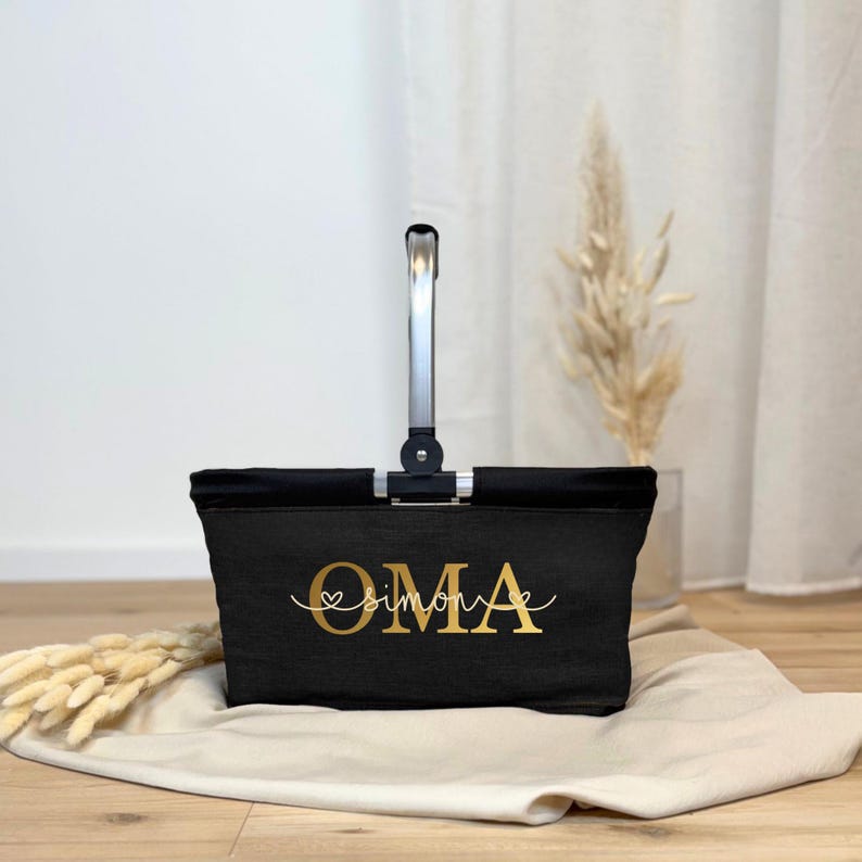 May include: A black tote bag with a silver handle and the word "OMA" in gold script. Below "OMA" is the name "simone" in a cursive font. The bag is sitting on a beige cloth.
