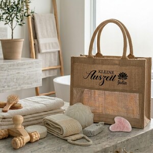 May include: A natural jute tote bag with handles, featuring the text "Kleine Auszeit Julia" and a lotus flower graphic. The bag is displayed with spa and bath products, including towels, a wooden massager, and a pink gua sha tool.