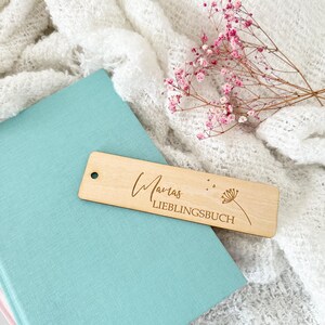 Könnte beinhalten: Ein Holz-Lesezeichen mit dem Text "Marias Lieblingsbuch" und einer kleinen Blumenillustration, das auf einem hellblauen Buch liegt. Das Lesezeichen ist hellbraun und hat ein Loch oben. Rosa Blüten und weißer Stoff sind im Hintergrund.