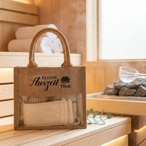May include: A natural burlap tote bag with a clear front panel, containing spa accessories. The bag has a jute handle and features the words "Kleine Auszeit Olivia" in black script and a lotus flower graphic. The background shows a sauna setting.