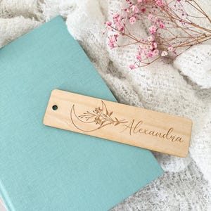 May include: A wooden bookmark with a crescent moon and floral design. The bookmark is engraved with the name "Alexandra".